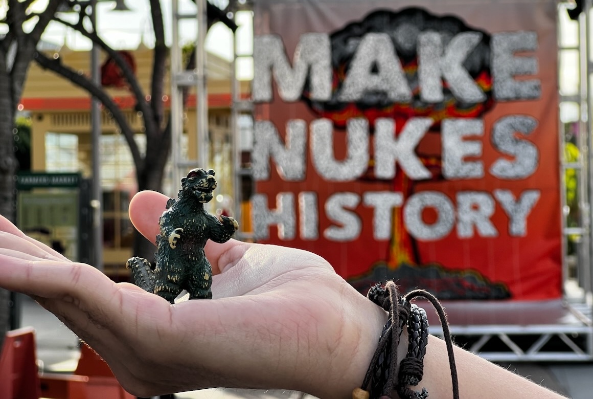 Hand holding small Godzilla toy in front of a large sign that reads Make Nukes History